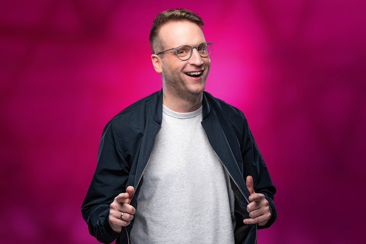 Benny Stark Profilbild Benny Stark zeigt mit zwei Fingern auf das Publikum vor rosanem Hintergrund