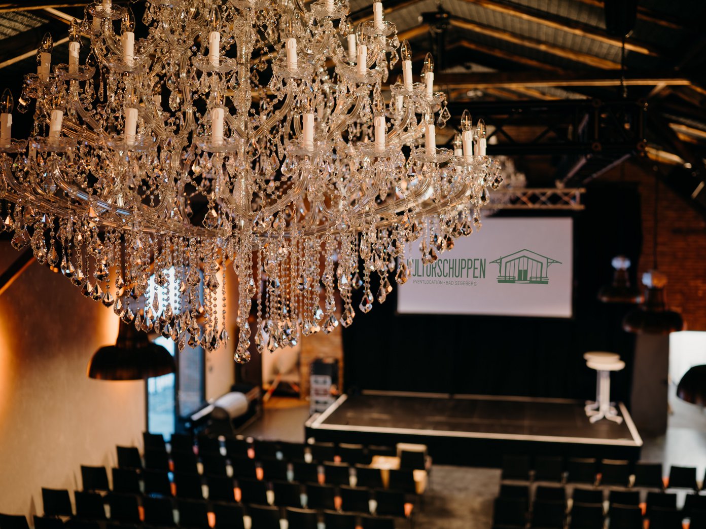 Kronleuchter Bestuhlung Kronleuchter vor Reihenbestuhlung, Leinwand mit Schriftzug "Kulturschuppen"
