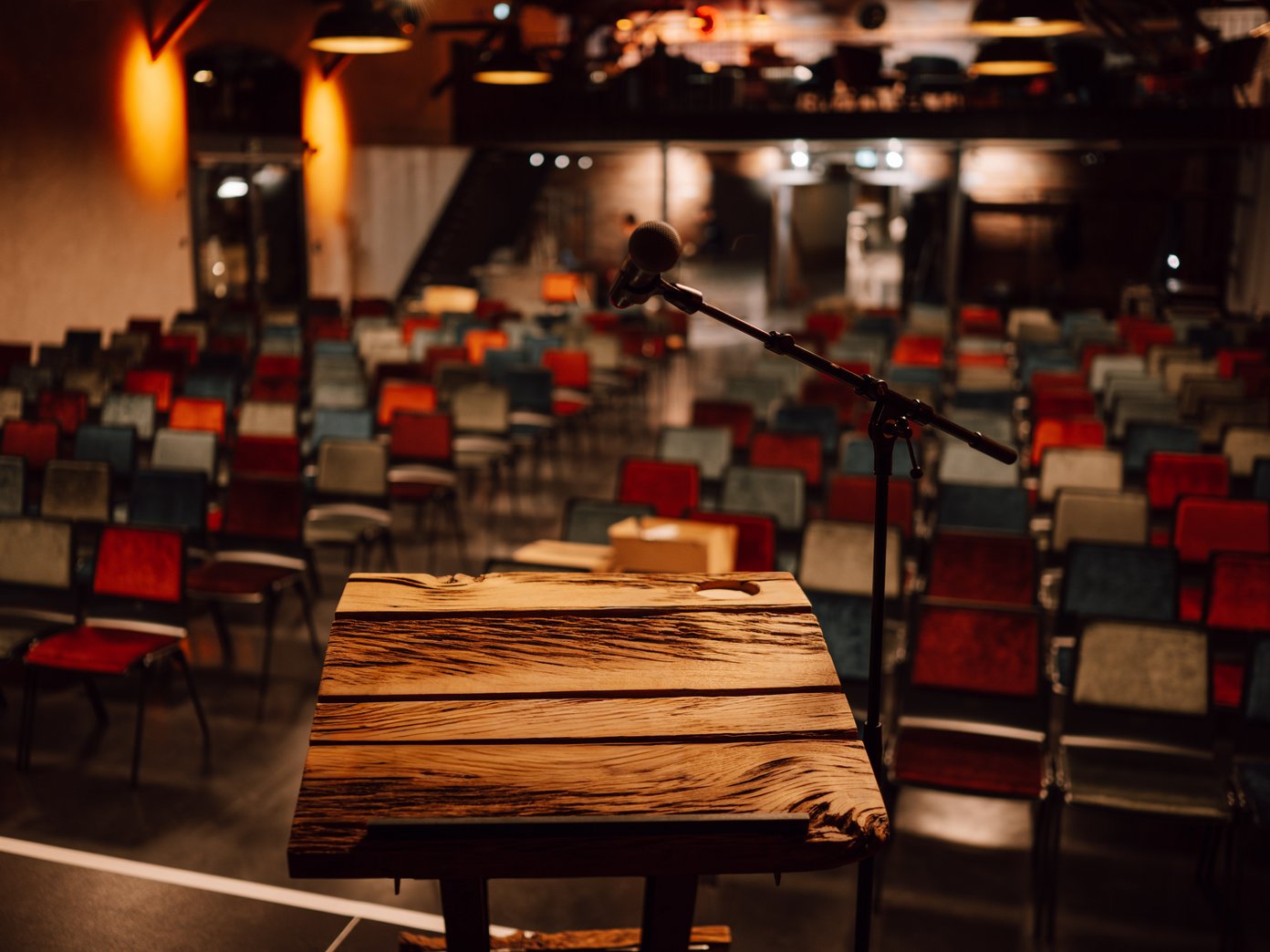 Vordergrund ein Holzpult vor einer Reihenbestuhlung mit bunten Stühlen