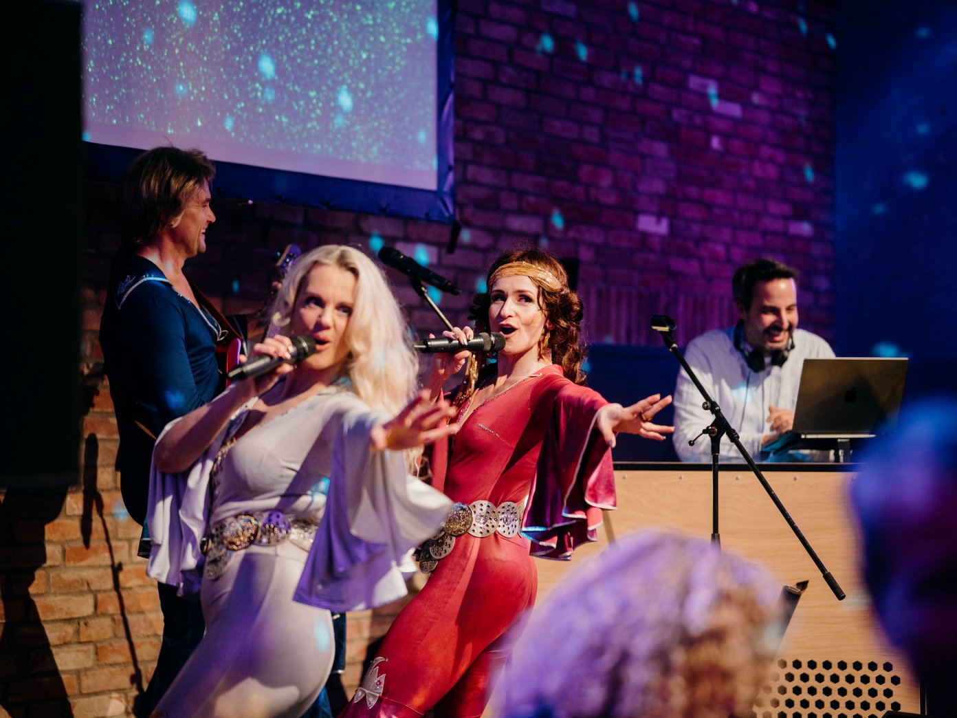 ABBA tanzend zwei weiblich aussehende Personen auf einer Bühe am singen, im Hintergrund ein DJ am DJ- Pult